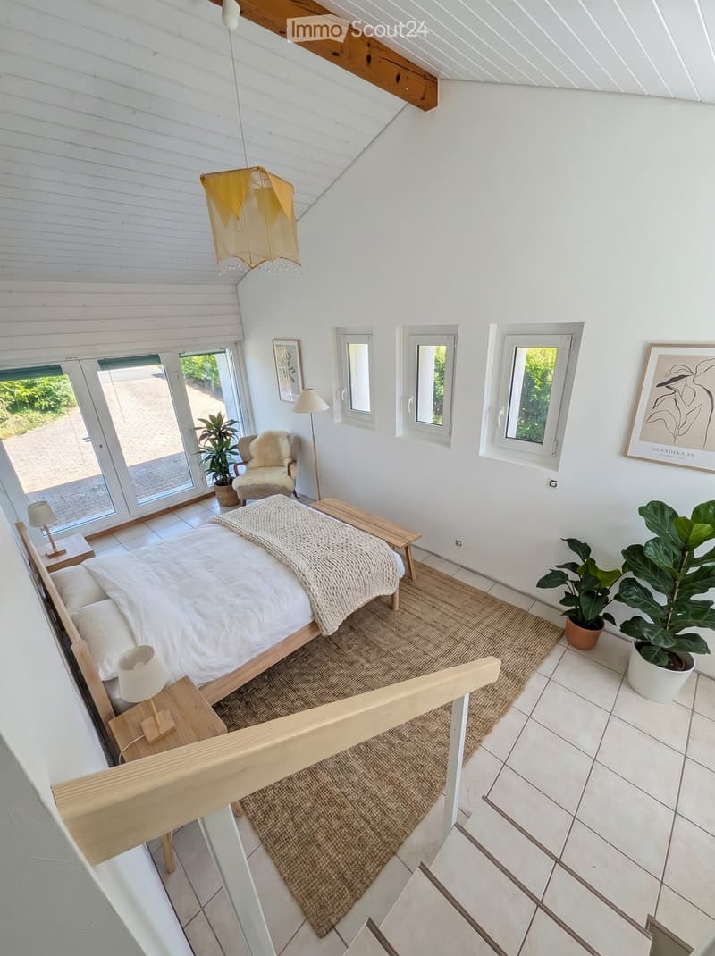 Bright, modern bedroom with white walls, wooden bed frame, nightstands, lamps, a cozy chair, a bench, a large potted plant, and artwork on the walls.