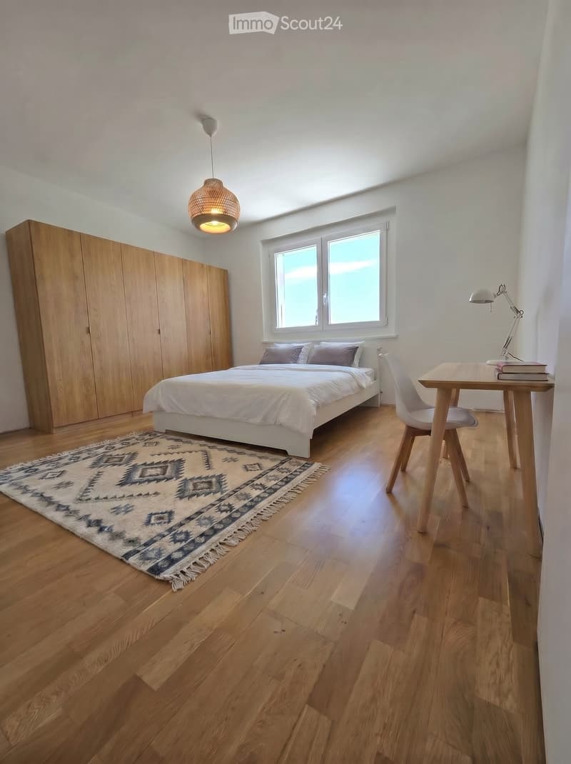 Modern bedroom with wooden flooring, a large bed with white bedding, a wooden wardrobe, a desk with a chair, a hanging pendant light, and a patterned rug.