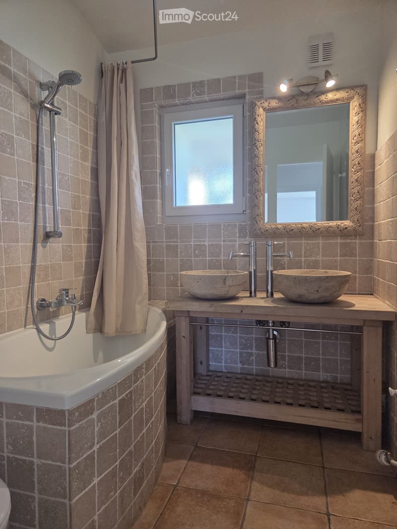 Bathroom with a bathtub, shower head, and a large mirror above a window. The room features tiled walls and floor, with a shower curtain partially drawn.