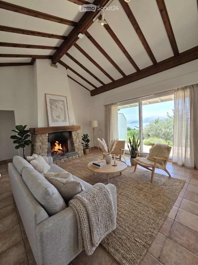 Living room with exposed wooden beams, fireplace, sofa, coffee table, and large windows offering a scenic view.