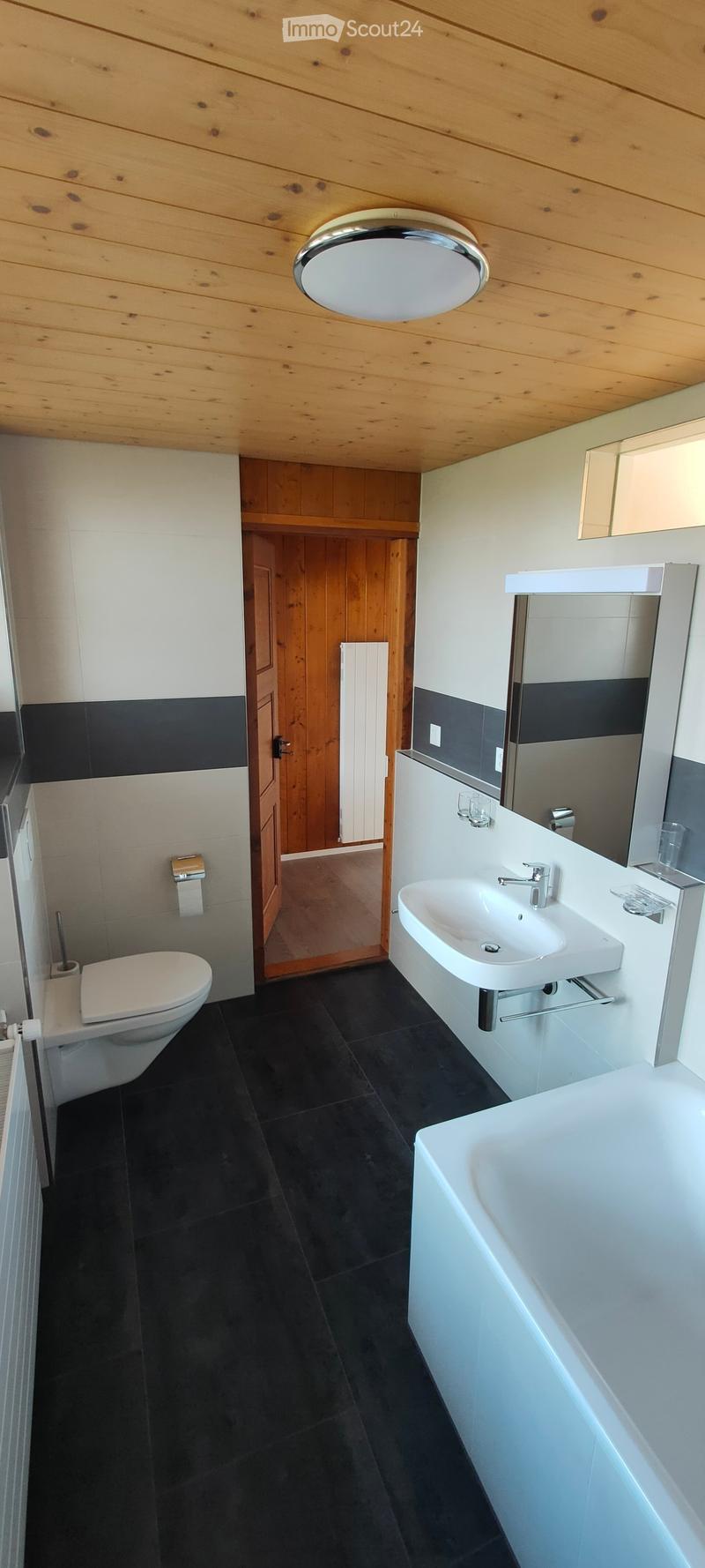 Bathroom with white bathtub, sink, toilet, wooden ceiling, black tiles, mirror, radiator