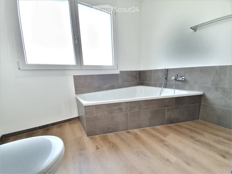 Modern bathroom with a bathtub, toilet, wooden flooring, and grey tiled walls.