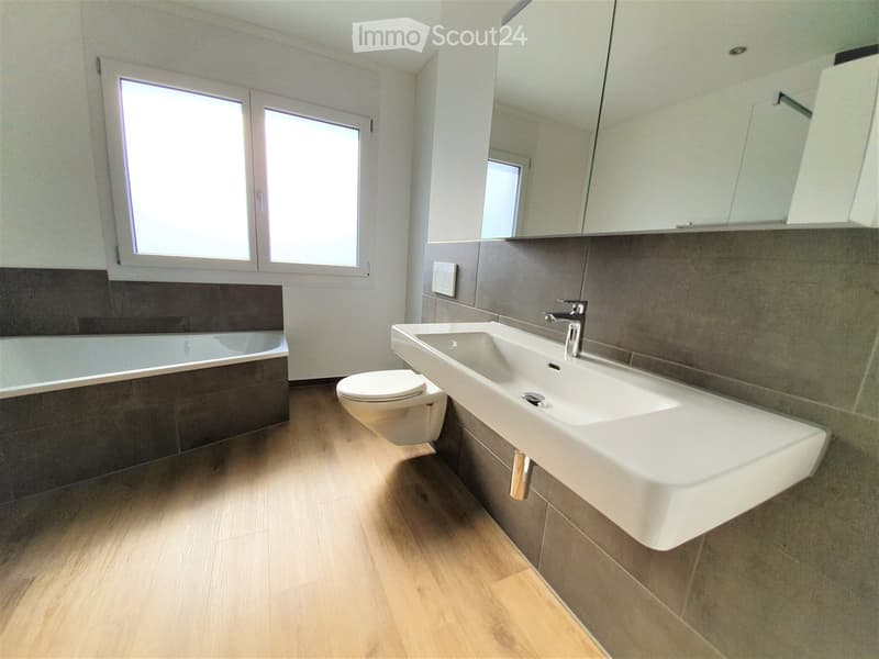Modern bathroom with a large sink, toilet, bathtub, and mirrored cabinet. The space features wooden flooring and large windows providing natural light.