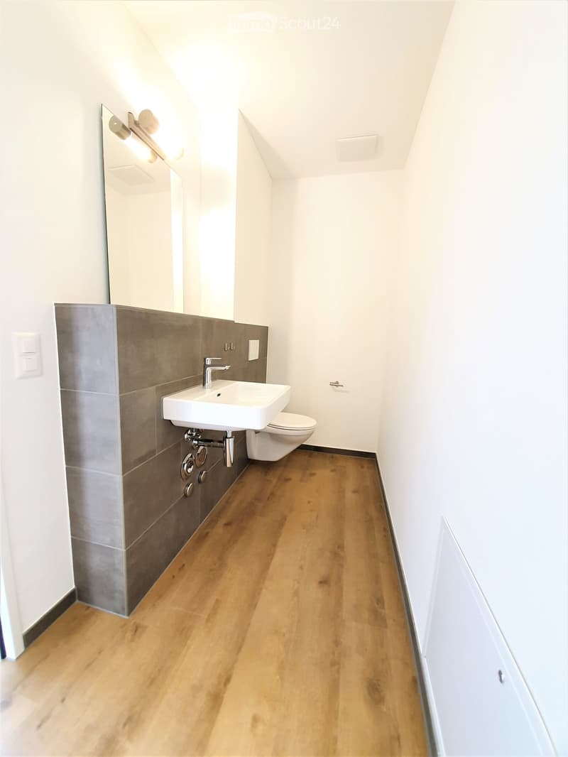 Modern bathroom with wooden flooring, white walls, a white sink, a toilet, and a large mirror with built-in lighting.