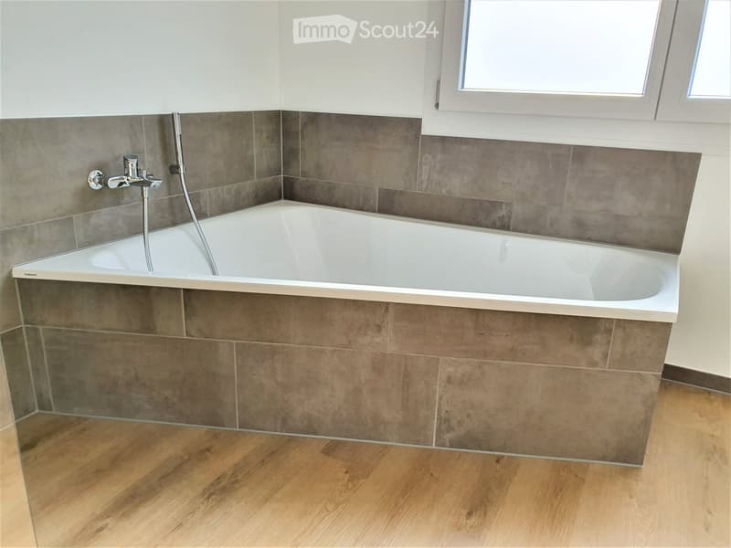 Modern bathroom with a white bathtub, grey tiled walls, and a wooden floor.