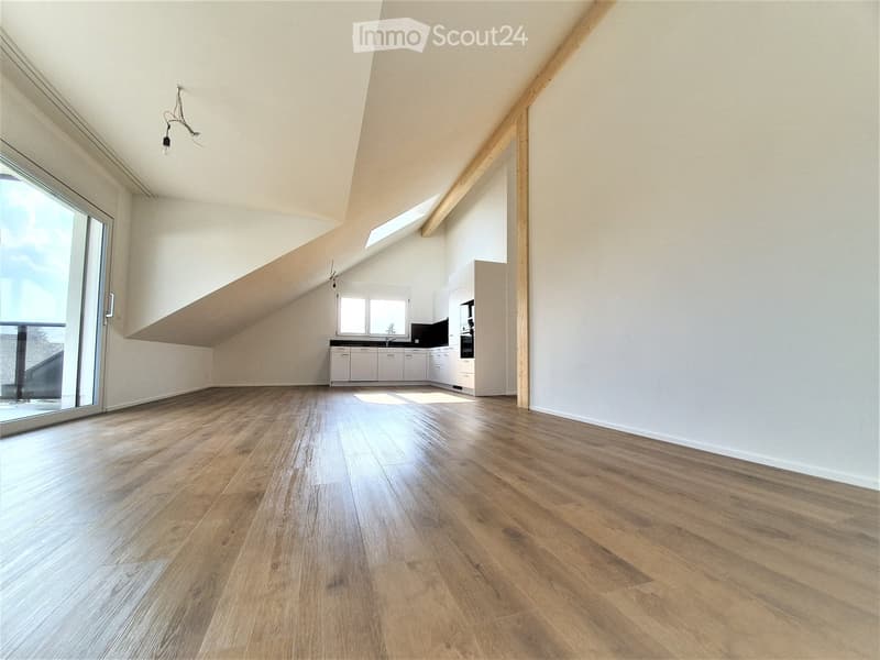 An unfurnished room with wooden flooring, white walls, and a small kitchen area in the background. The room features large windows and a sliding glass door leading to an outdoor area.