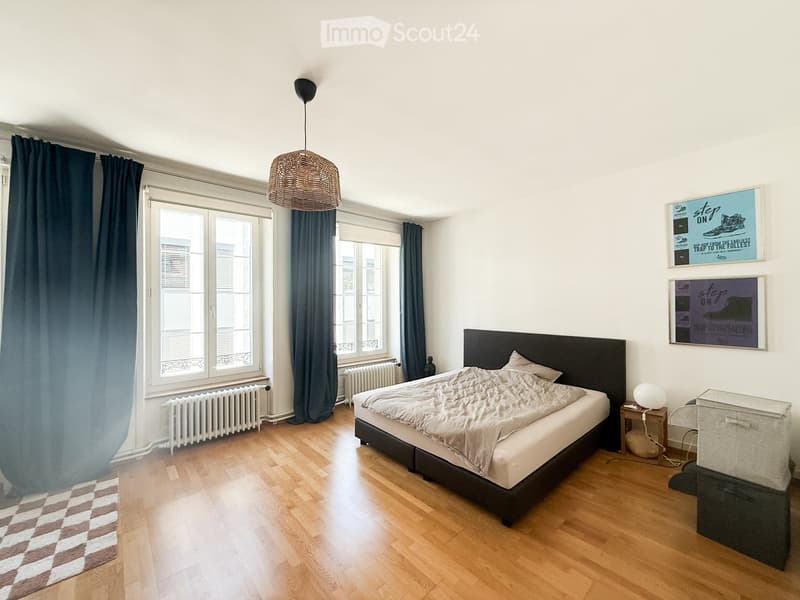 A modern bedroom with wooden flooring, a large bed with a dark headboard, a nightstand with a lamp, and two framed pictures on the wall. The room features large windows with dark curtains and a radiator below them.