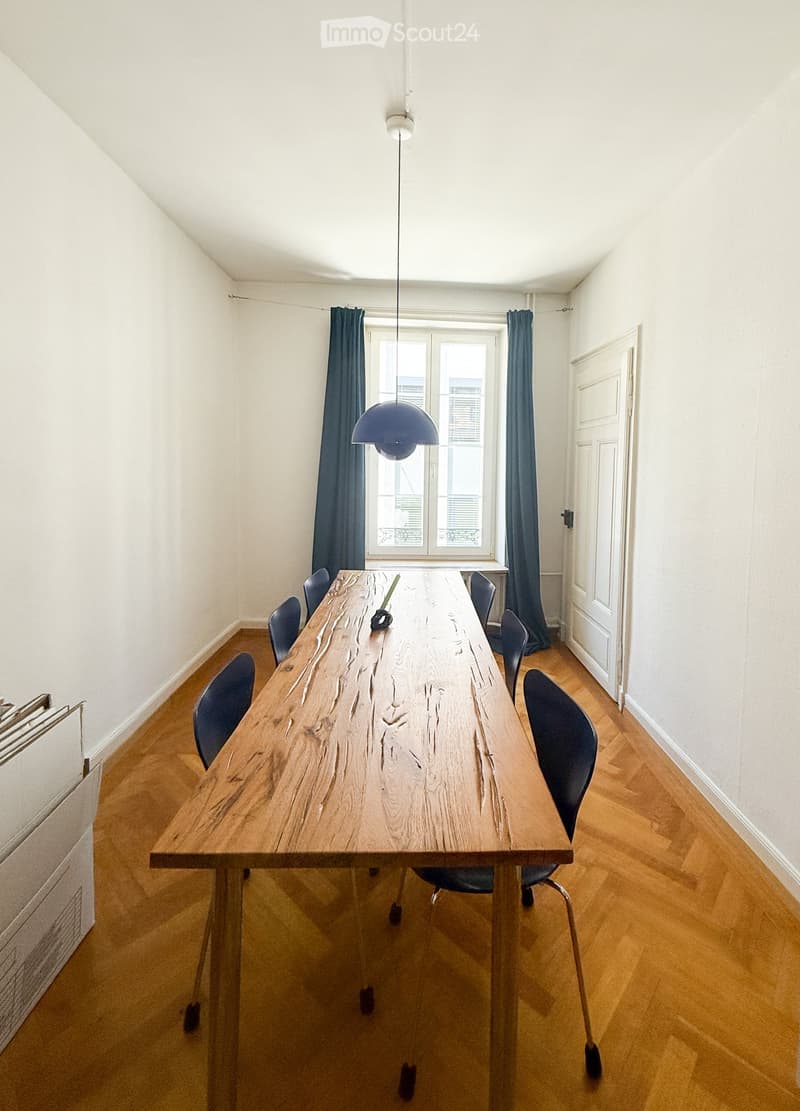 Bright room with wooden floor, large wooden table surrounded by chairs, blue curtains, and a hanging lamp.