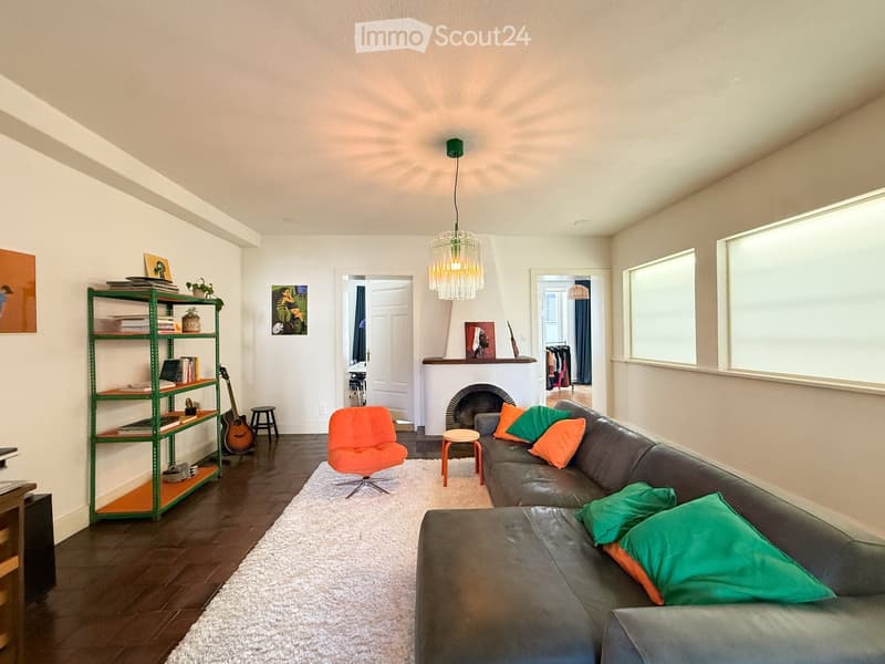 Modern living room with dark hardwood floors, white walls, a fireplace, a large sectional sofa with colorful pillows, a bright orange chair, a green metal shelving unit with books and a laptop, a guitar, and a hanging chandelier.