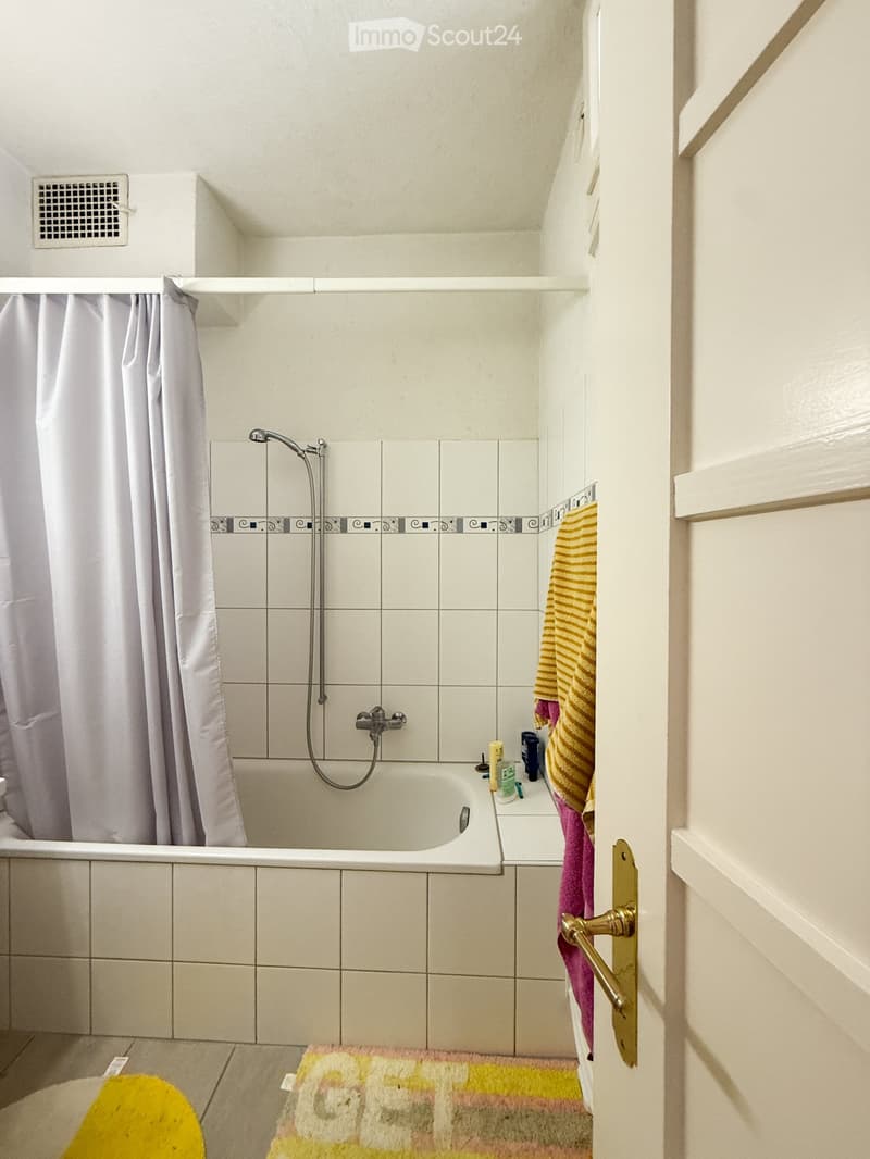 Bathroom with a white bathtub, tiled walls, a shower curtain, a towel hanging on the side, and various bathroom accessories on the counter.