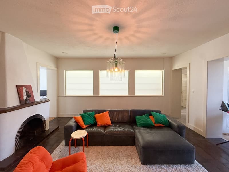 Modern living room with a dark leather sectional sofa, colorful throw pillows, a small wooden stool, an orange armchair, a fireplace with a painting above it, and a chandelier hanging from the ceiling.