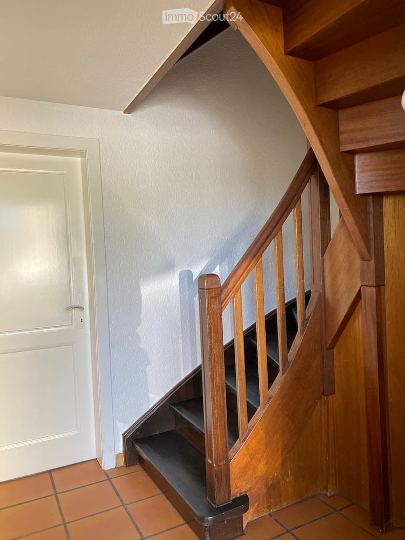 Wooden staircase with a handrail, leading up to a landing area. The steps are carpeted, and the walls are painted white. A white door is visible on the left side.