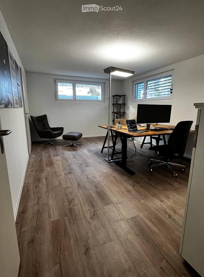 A modern, well-lit office space with wooden flooring, a large desk, a laptop, a screen, a chair, a stool, a floor lamp, and a small shelf.
