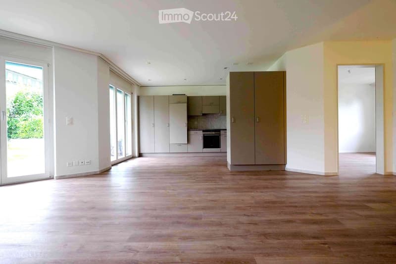 Empty room with wooden flooring, white walls, large windows, and a kitchen area with cabinets and appliances visible in the background.