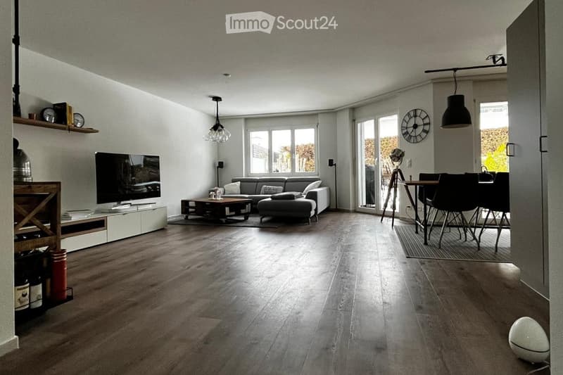 Modern living room with grey sofa, coffee table, ottoman, TV stand, and dining area with table and chairs. Features wooden flooring, large windows, and pendant lighting.