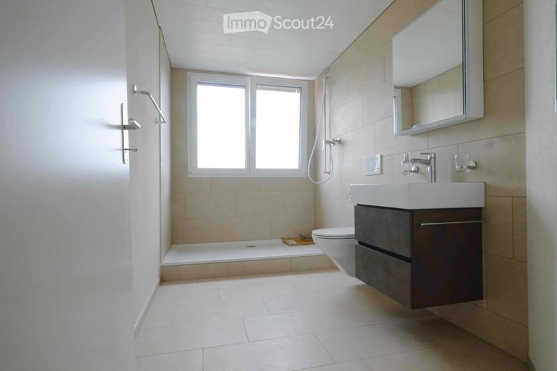 Modern bathroom with a bathtub, toilet, sink, mirror, and shower head. Light-colored tiles and a window provide natural light.