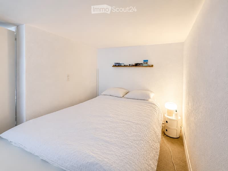 Minimalist bedroom with a white bed, nightstand, lamp, and a small shelf with photos and decorative items.