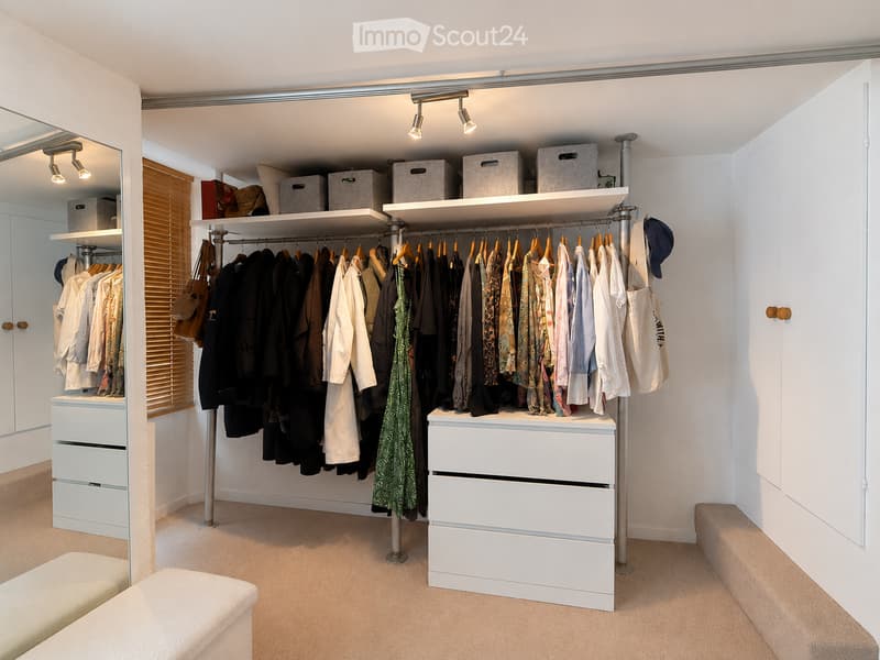 A well-organized walk-in closet with white built-in wardrobes, multiple shelves, and drawers. Clothing is neatly hung on rods, and various items such as handbags and umbrellas are visible.