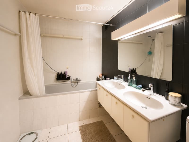 Modern bathroom with a bathtub, double sink, large mirror, and a shower area with a curtain. Includes a bathroom scale and a rug on the floor.