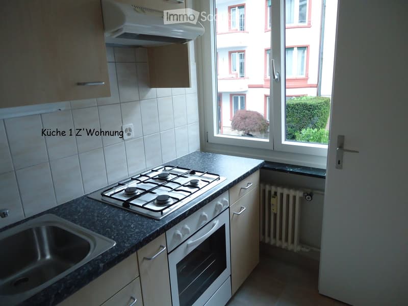 Compact kitchen with a gas stove, oven, sink, and cabinets, featuring a window with a view of a building and greenery outside.