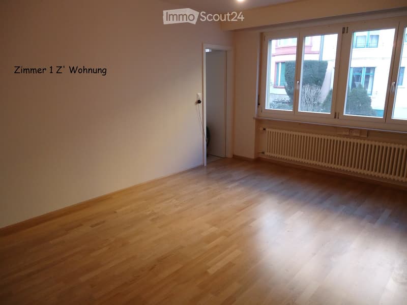 Empty room with light wooden flooring, white walls, large windows, and a radiator below the windows. No furniture or personal items are present.