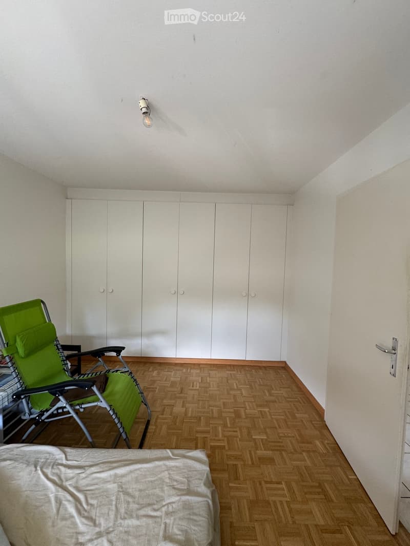 A small, furnished room with wooden herringbone flooring, white built-in wardrobes, a green folding chair, and a bed with a white cover.