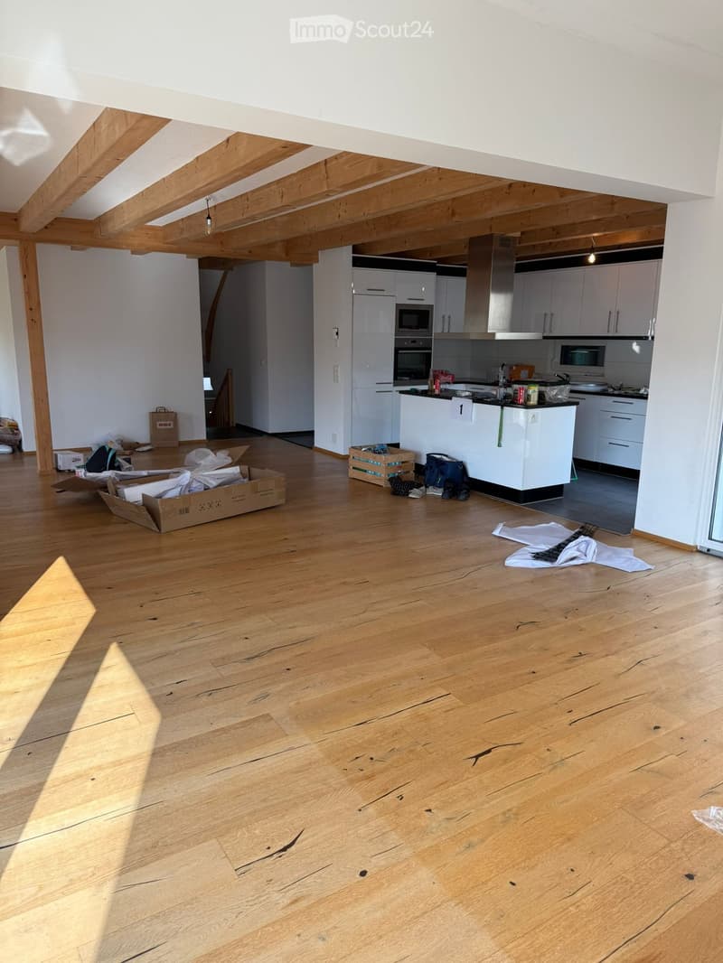 Empty room with wooden flooring, white walls, and exposed wooden beams on the ceiling. Kitchen area visible in the background with cabinets and appliances.