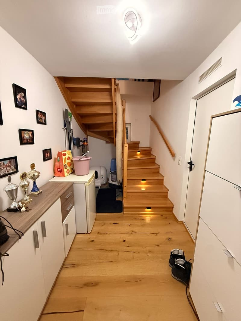 Wooden staircase with built-in lighting, white walls, and a small shelf with trophies and a pink bucket. The hallway has a wooden floor and white cabinets on either side.