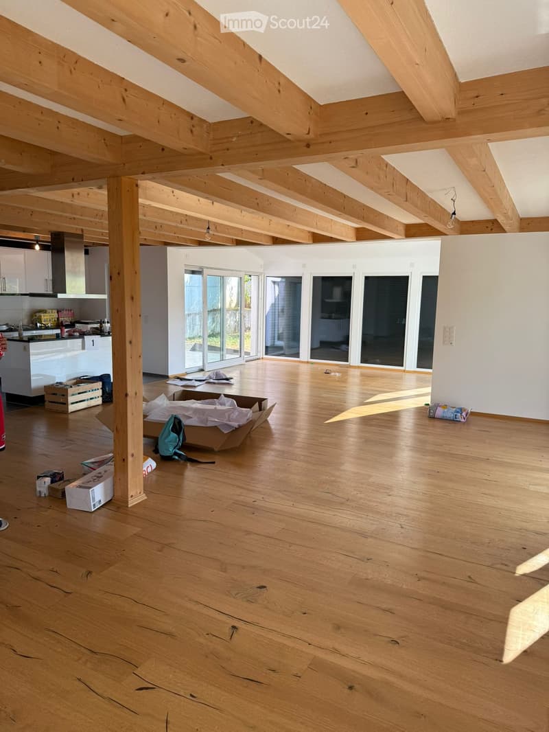 Open, unfurnished room with wooden floors, exposed wooden beams, and large windows. Some boxes and items are scattered on the floor.