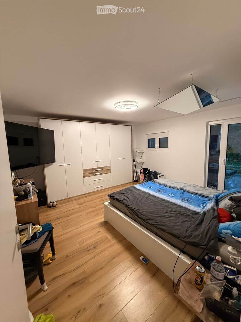 Modern bedroom with wooden flooring, large bed with blue and grey bedding, white wardrobe with drawers, a chair with clothes, a small table with items, and a large screen on the wall.