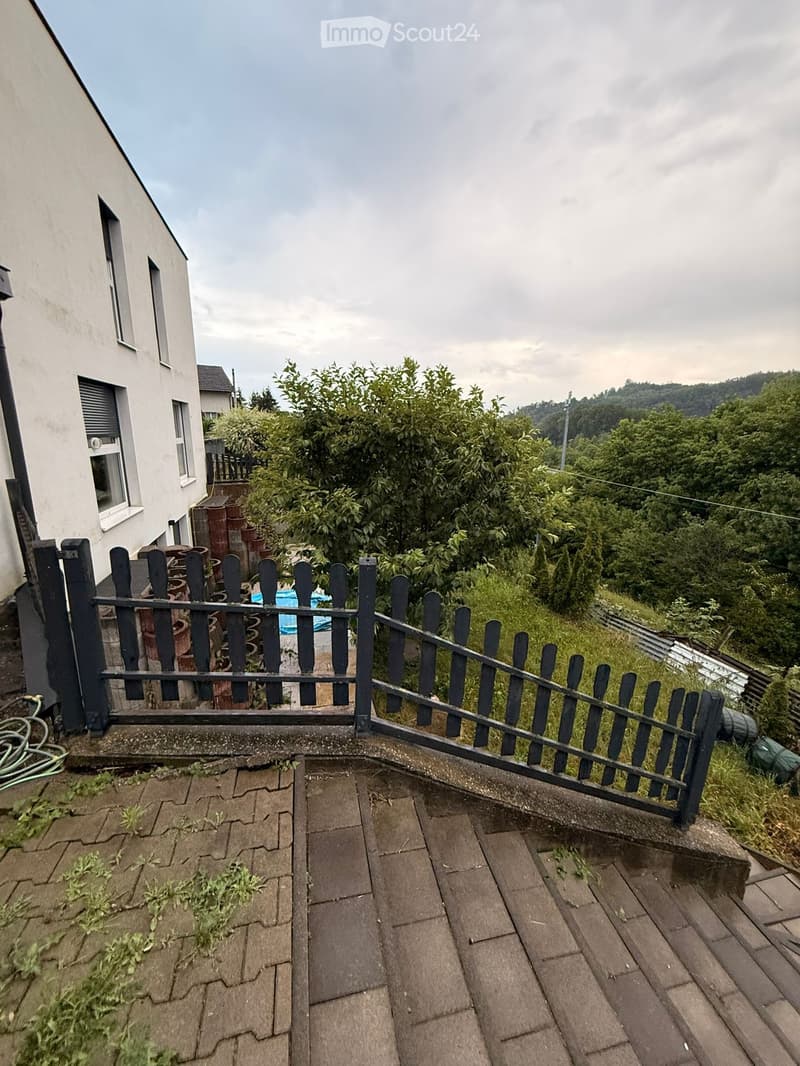 Balcony with brick tiles, metal fence, and view of greenery and a neighboring house.
