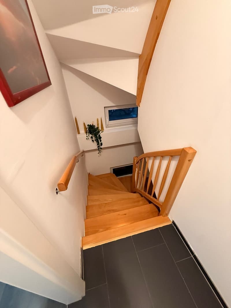 Wooden staircase with handrails, white walls, small window, and a small plant on the wall.