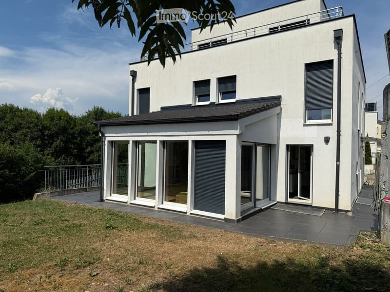 Modern two-story house with white exterior, large windows, and a raised ground floor. Features include a balcony on the upper level and a spacious patio area with glass doors leading to the interior.