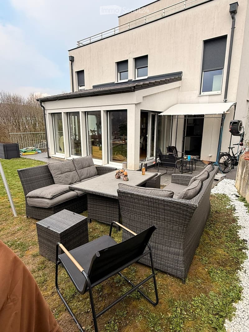 Outdoor patio area with wicker furniture, including a sofa, table, and chairs, near a modern house with large glass doors and a covered patio area.