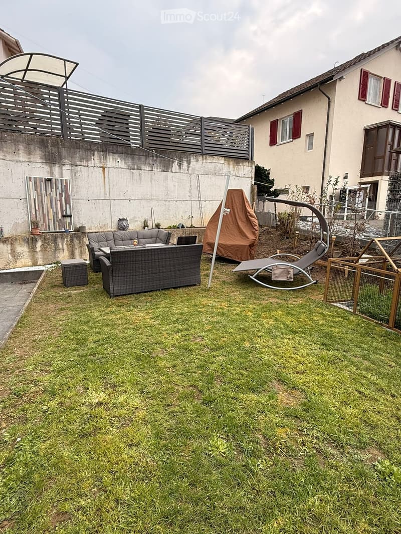 Garden with a sofa, table, chairs, a swing chair, and a covered grill. The area is enclosed by a fence and has a concrete wall with a mirror and small plants.