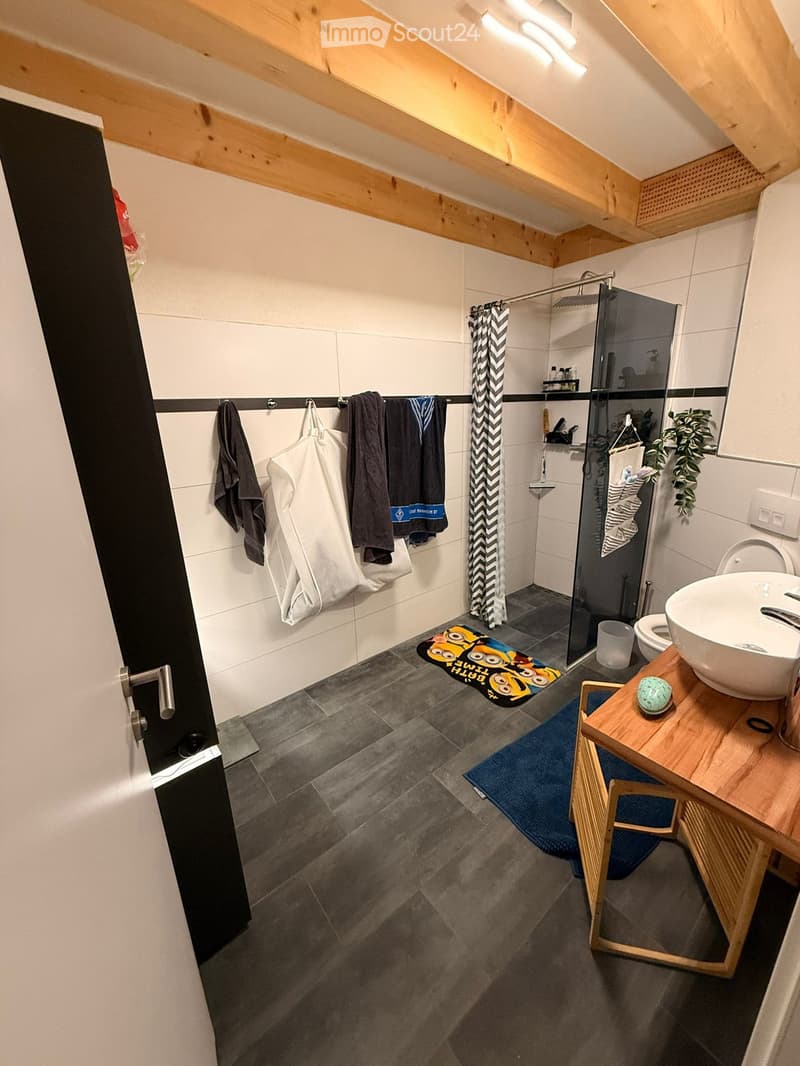 Modern bathroom with a glass-enclosed shower, a sink with a wooden cabinet, a toilet, and a towel rack with towels. The floor is tiled, and there is a decorative rug in front of the sink.