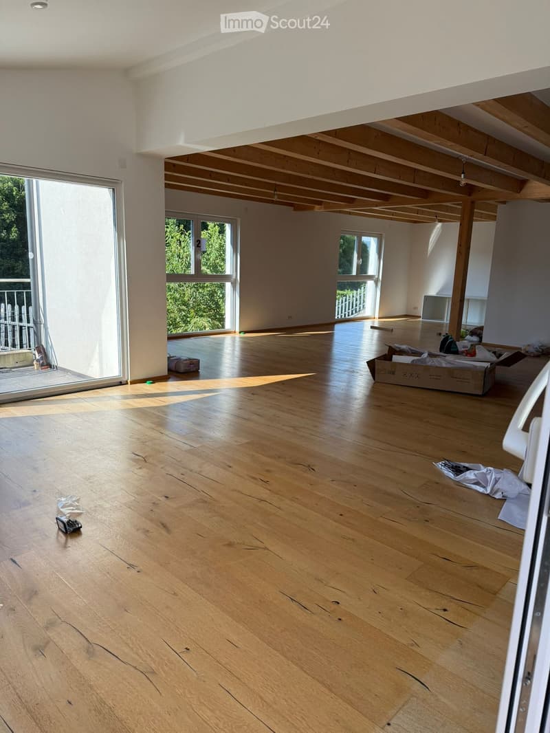 Empty room with wooden flooring, white walls, and exposed wooden beams on the ceiling. Large windows and sliding doors lead to a balcony area with outdoor greenery visible.
