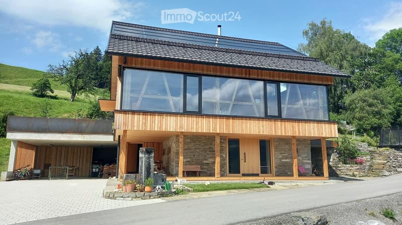 Modern two-story house with wooden and stone exterior, large windows, raised ground floor, garage, and outdoor seating area.