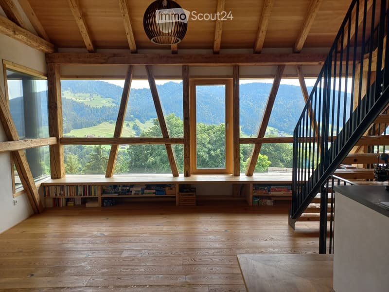 Bright, open living space with wooden floors, large windows offering a scenic mountain view, a built-in bookshelf, and a modern staircase leading upwards.