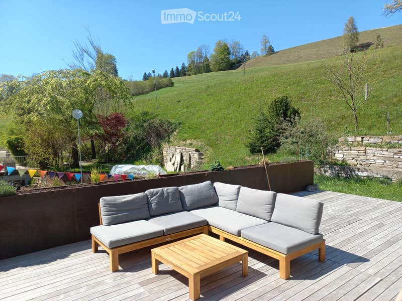 Wooden deck with grey cushioned sofa and coffee table, surrounded by greenery and a stone wall.