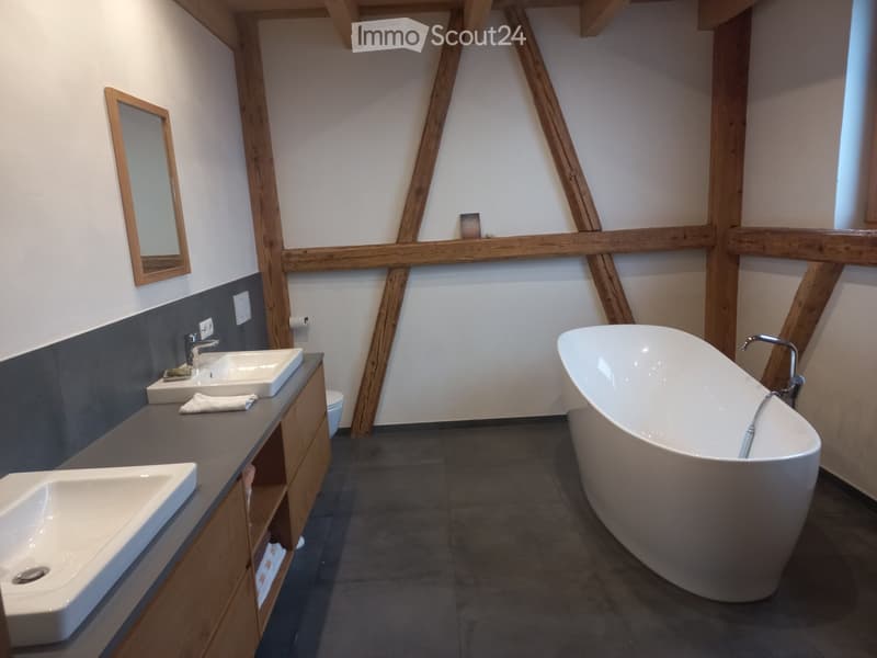 Modern bathroom with a freestanding bathtub, double sink vanity, and exposed wooden beams. Features a large mirror, sleek faucet, and a small decorative item on the shelf.