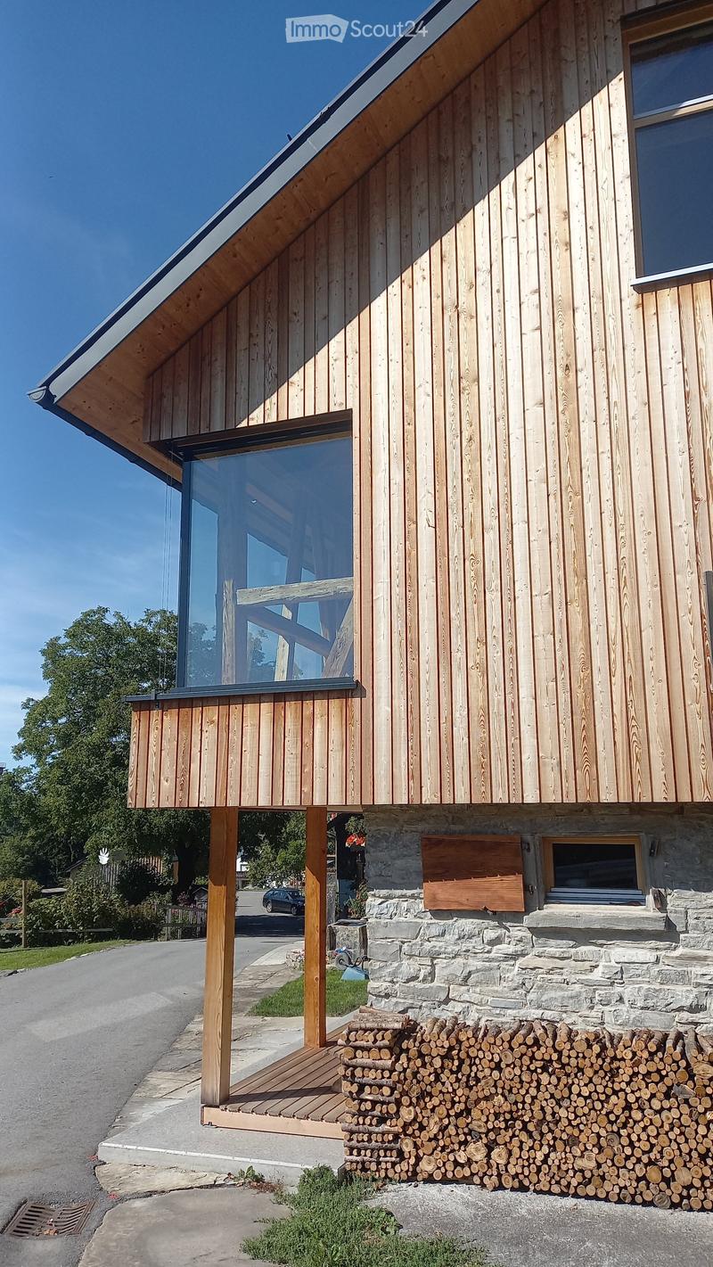 Modern two-story house with wooden siding, large glass windows, stone base, and a wooden deck with stacked firewood nearby.