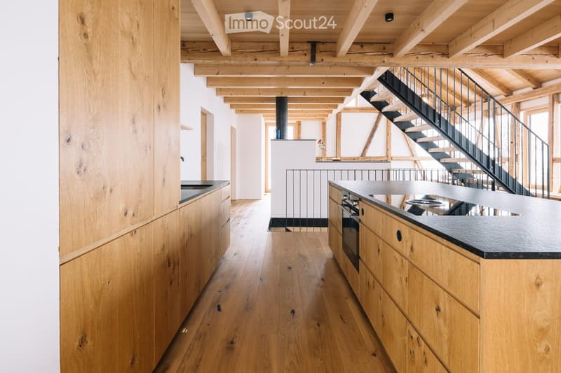 Modern kitchen with light wooden cabinets, a black countertop, and a visible stove. The space features exposed wooden beams on the ceiling and a staircase leading to an upper level.