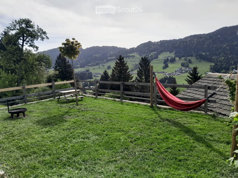 A grassy outdoor area with a wooden fence, a red hammock, a picnic table, and a scenic mountain view in the background.