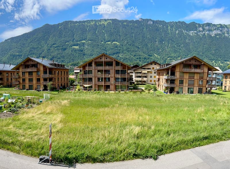 A cluster of multi-story wooden buildings with balconies, surrounded by a grassy field and set against a backdrop of a mountain range.