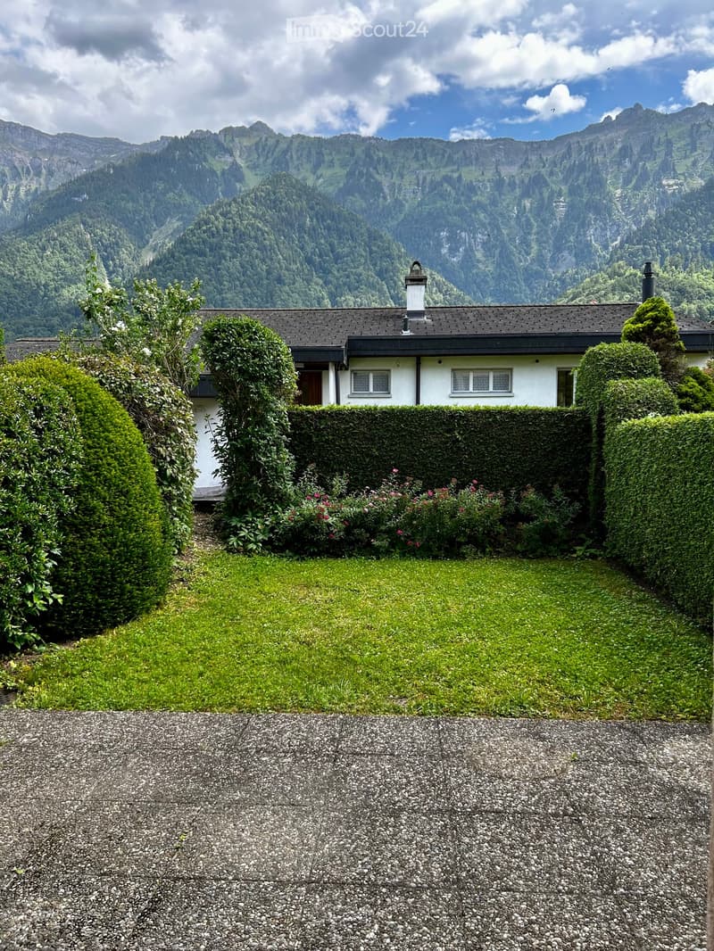 A single-story house with a dark roof and white walls, surrounded by a well-maintained garden with hedges, shrubs, and a gravel driveway, with a scenic mountain backdrop.