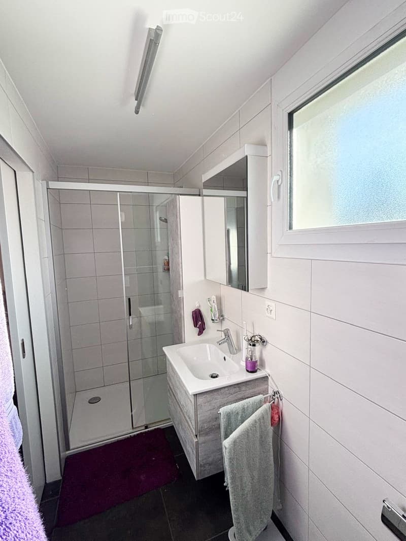 Modern bathroom with a white sink, mirror, shower, and towels. Features a sleek design with a window and built-in storage.