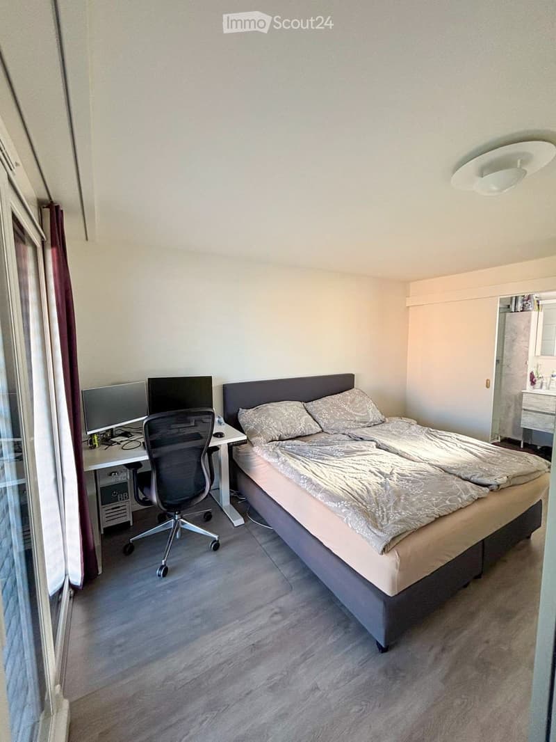 A modern bedroom with a double bed, desk with computer, office chair, and large window with curtains.