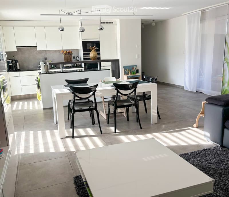 Open-plan living and dining area with modern white furniture, a dining table with chairs, a sofa, coffee table, and a kitchen area with cabinets and appliances.