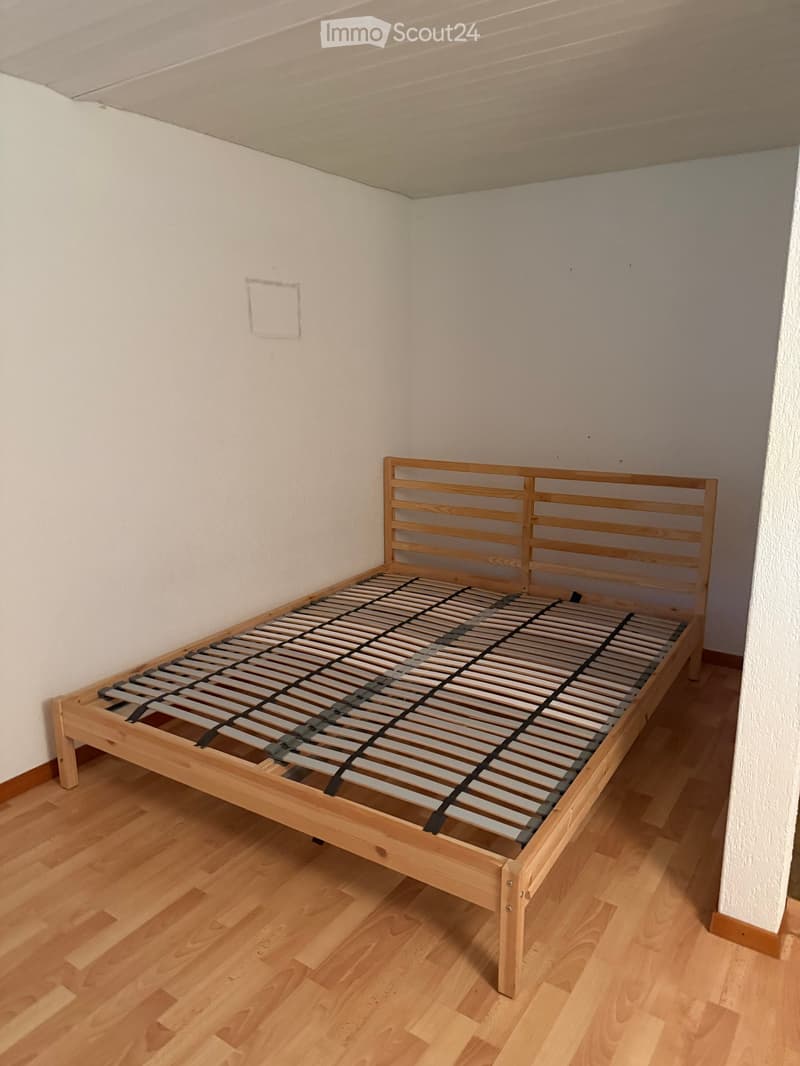 A wooden bed frame with a slatted mattress base is placed against a white wall in a room with light wooden flooring.